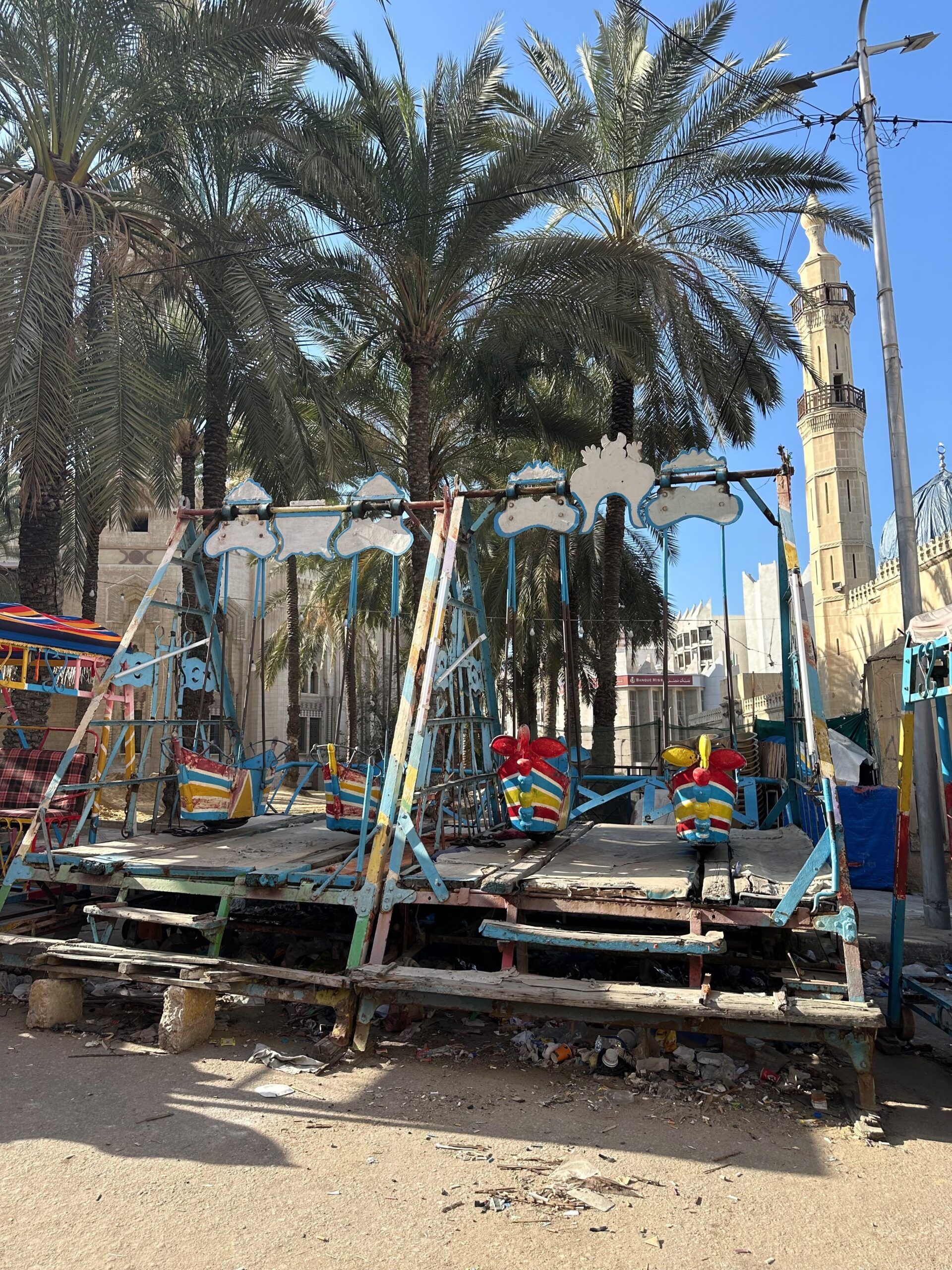 Abandoned colorful swing ride beneath palm trees near a mosque minaret in egypt