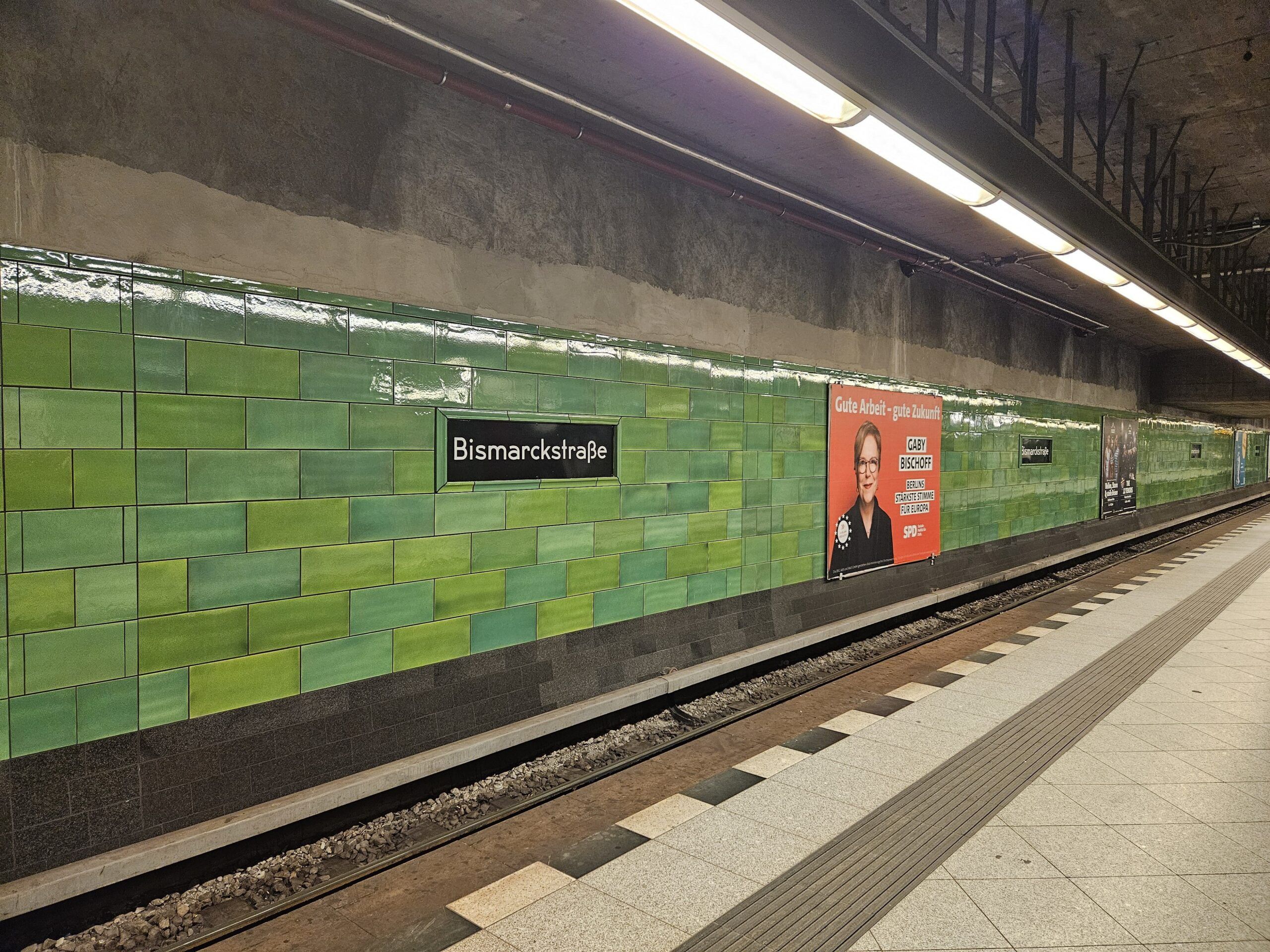 Peron stacji bismarckstraße w berlińskim metrze z zielonymi kafelkami i reklamą wyborczą.
