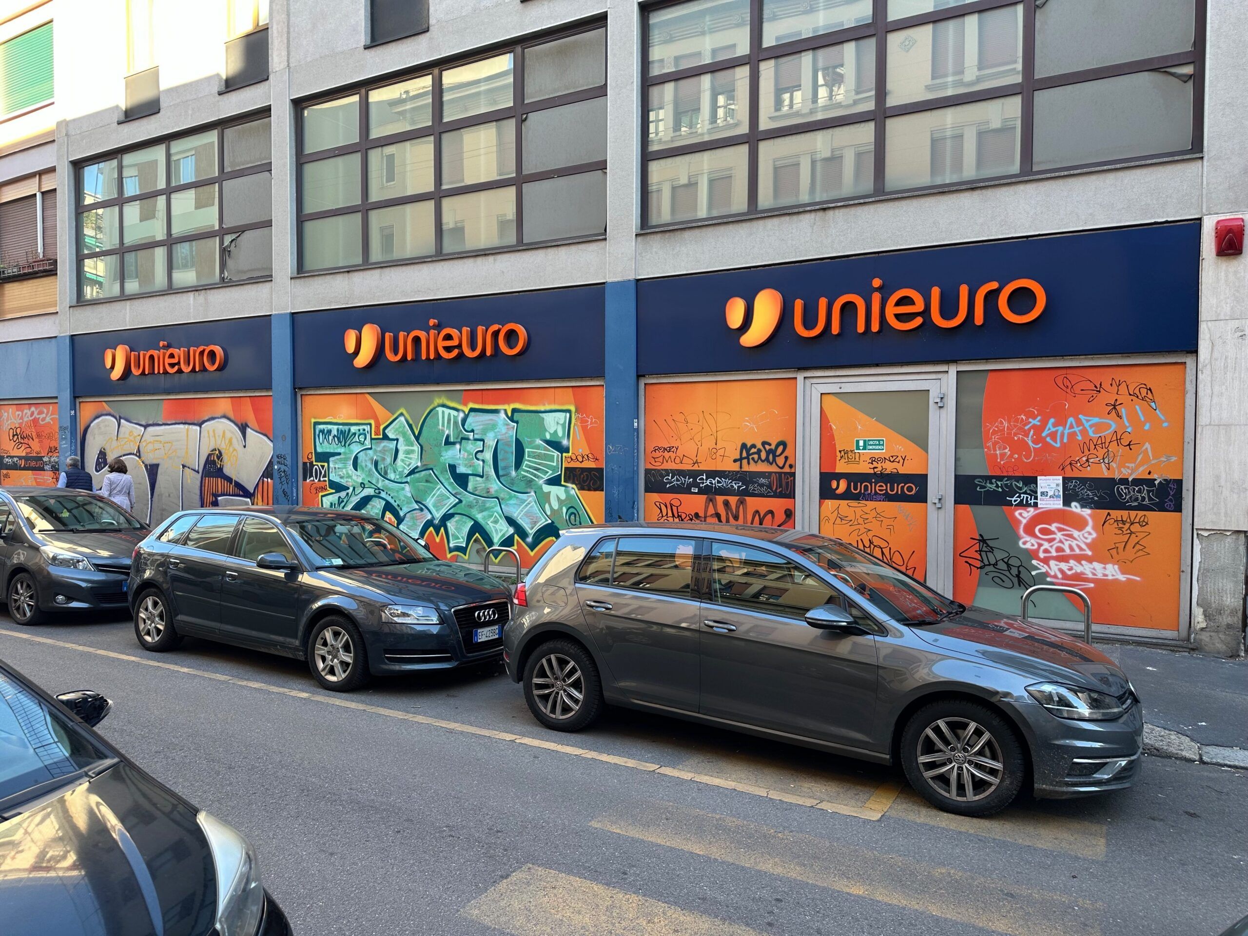 Zniszczona elewacja sklepu unieuro w mediolanie z graffiti i zaparkowanymi samochodami.