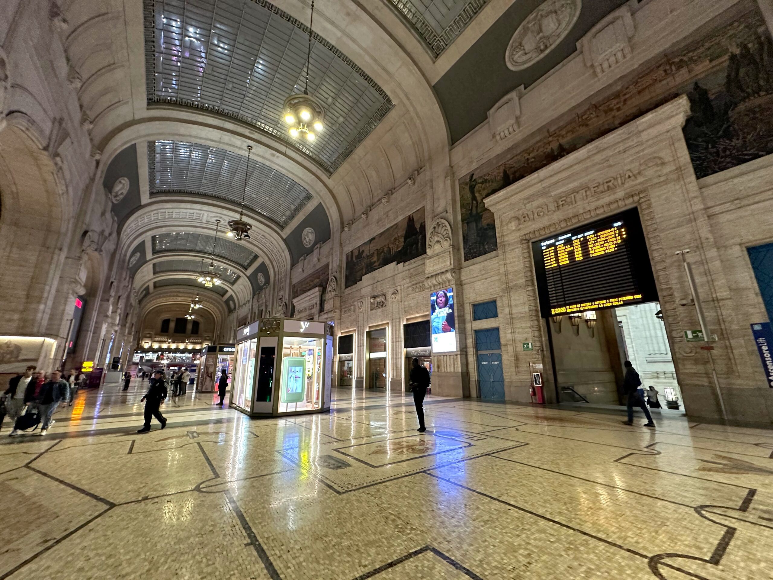 Wnętrze dworca milano centrale z wysokim sklepieniem, tablicą odjazdów i pojedynczymi podróżnymi.