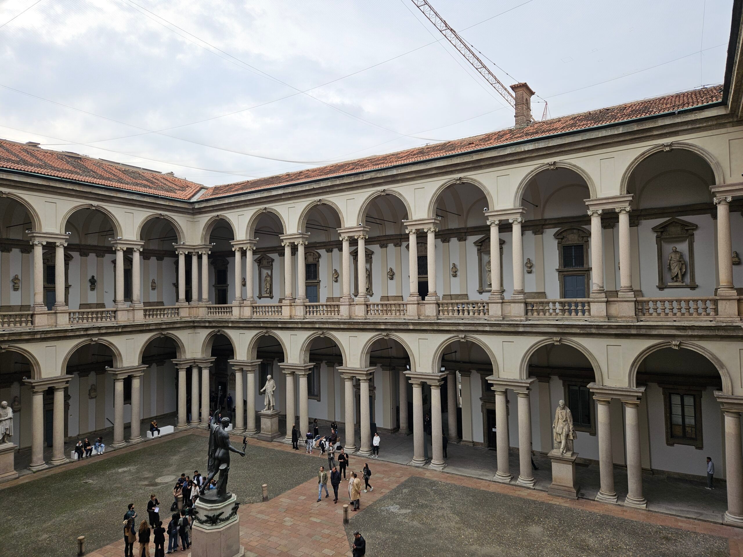 Dziedziniec pinacoteca di brera w mediolanie z arkadami i rzeźbami osób zwiedzających.