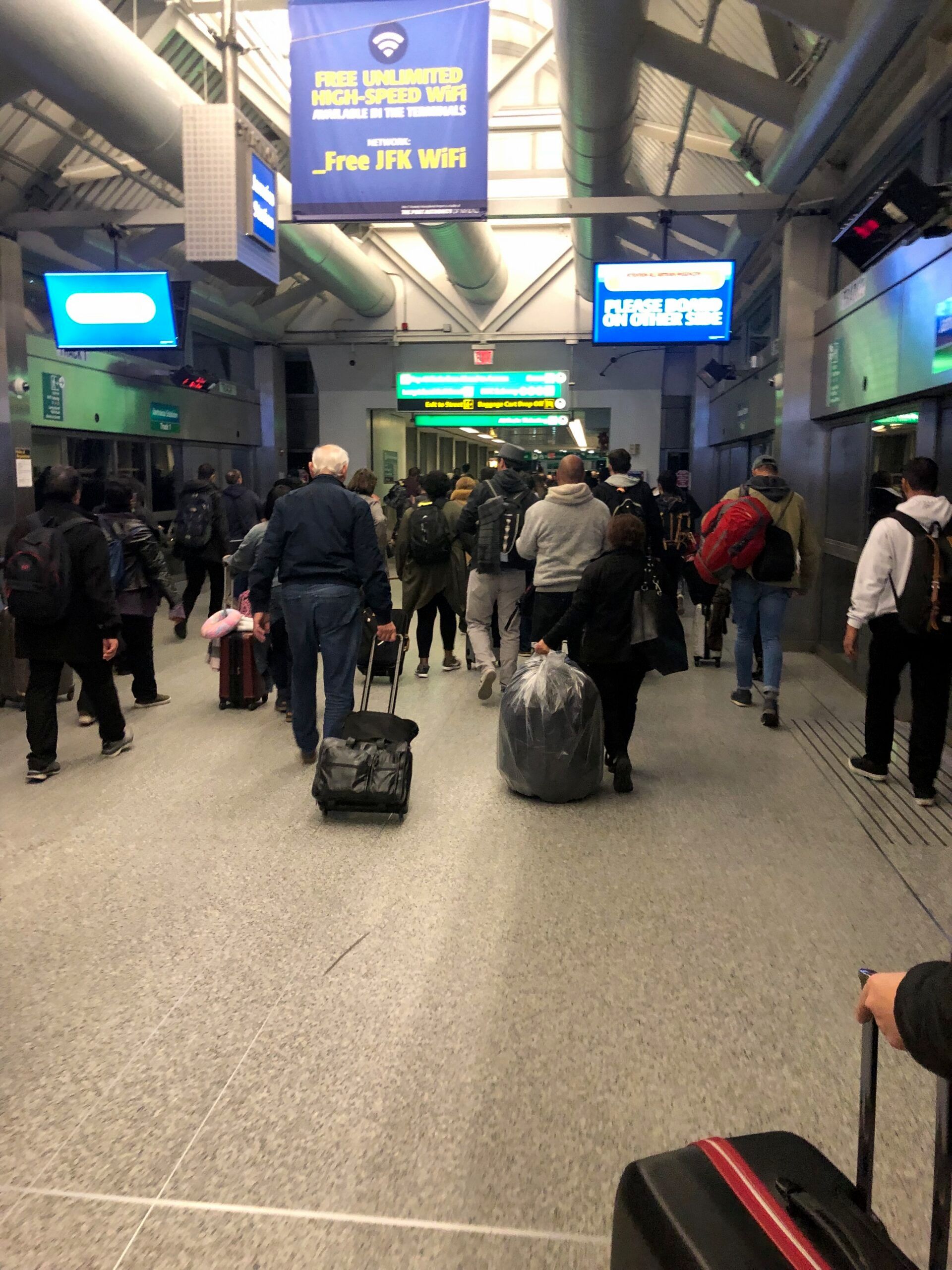 Crowds wait in a transit station near signs offering free jfk wifi, with suitcases and luggage carts.