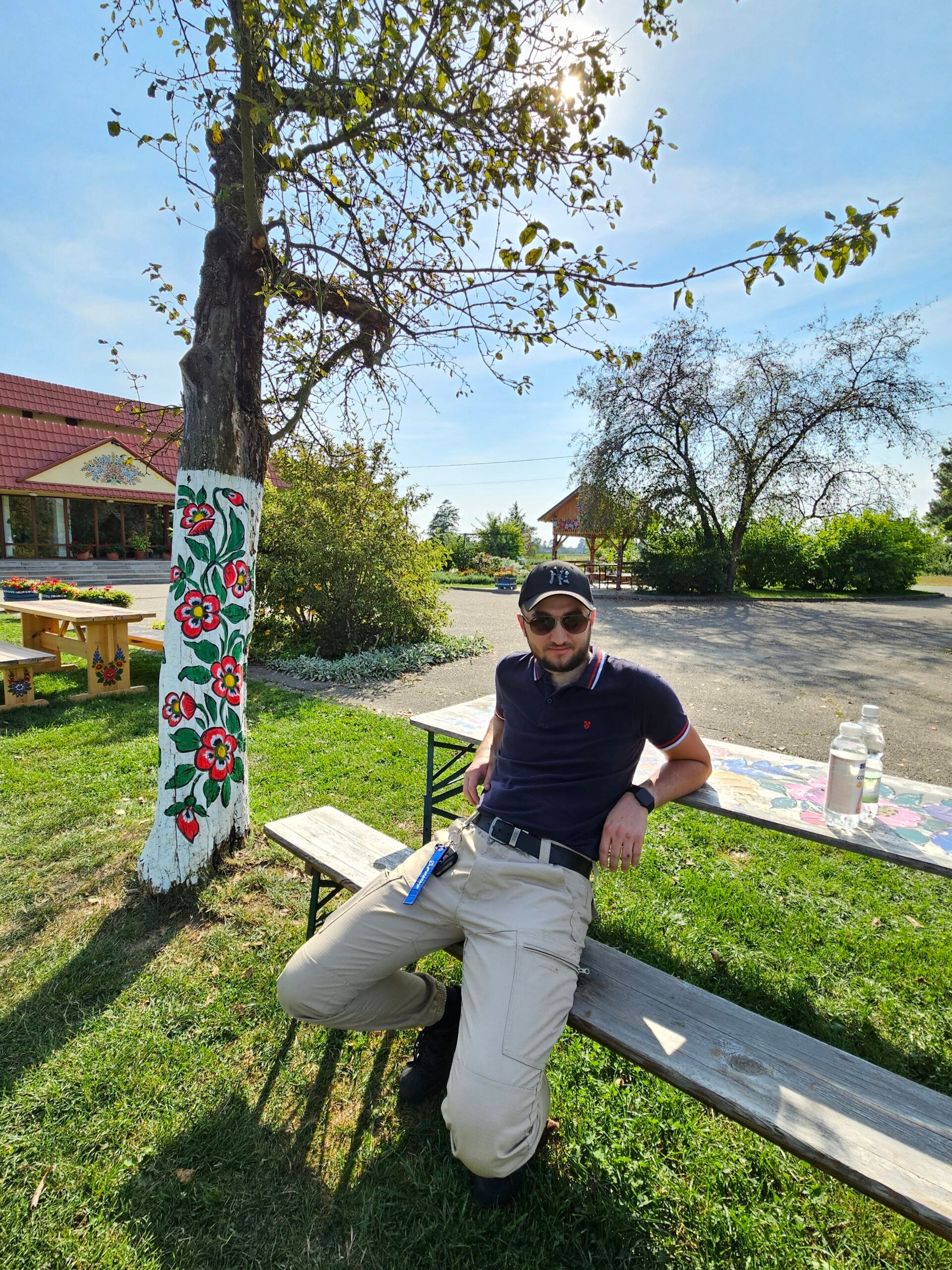 Zalipie women painters’ house: mężczyzna siedzi na ławce przy zdobionym kwiatami drzewie i budynku.