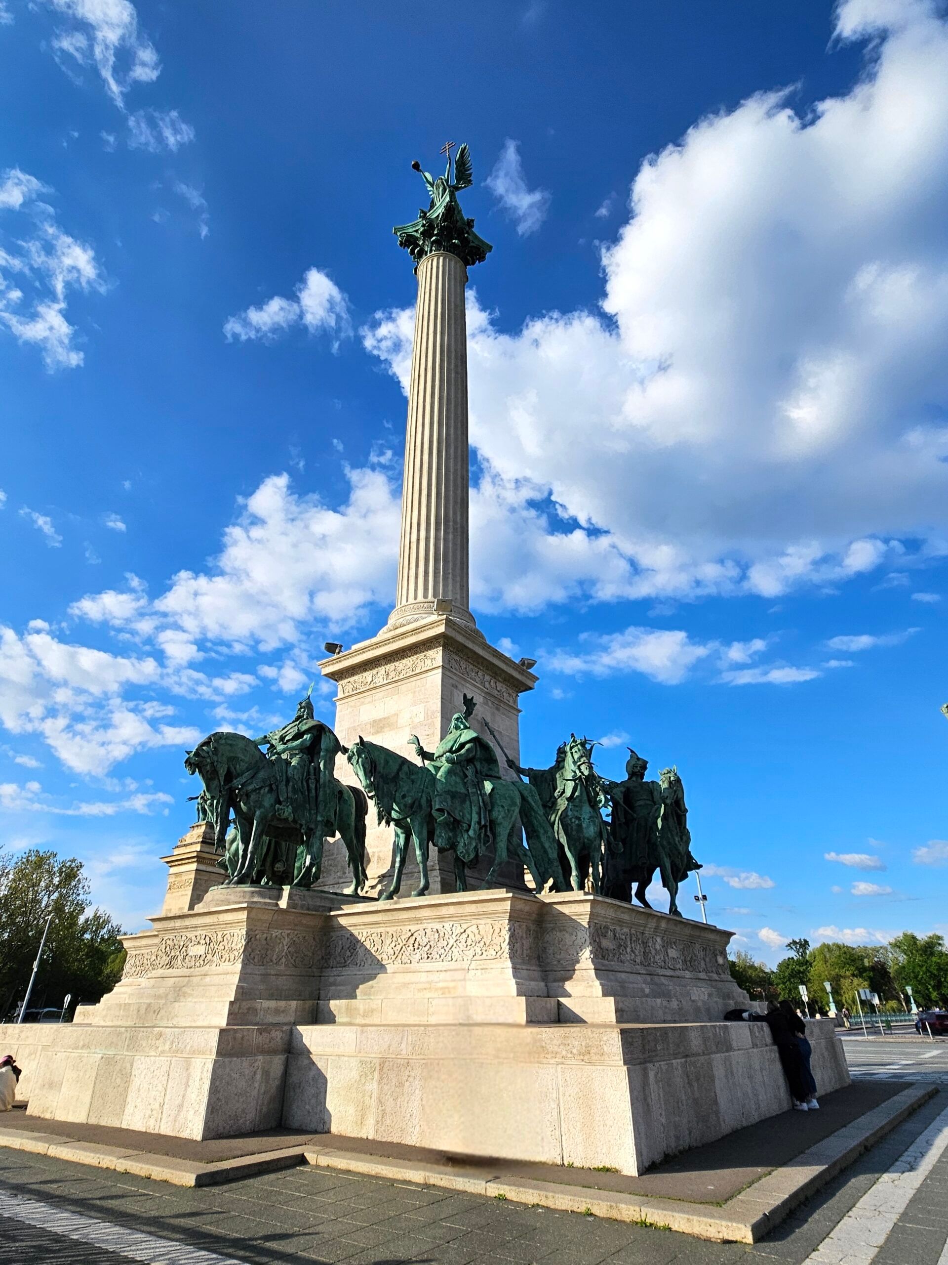 Plac bohaterów w budapeszcie z pomnikiem kolumnowym i brązowymi posągami jeźdźców.