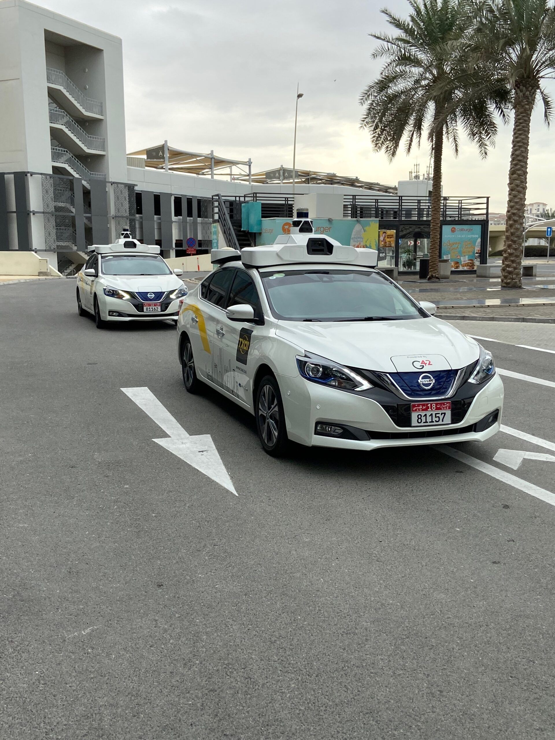 Autonomiczne taksówki txai w abu dhabi stoją na ulicy przy palmach i nowoczesnym budynku