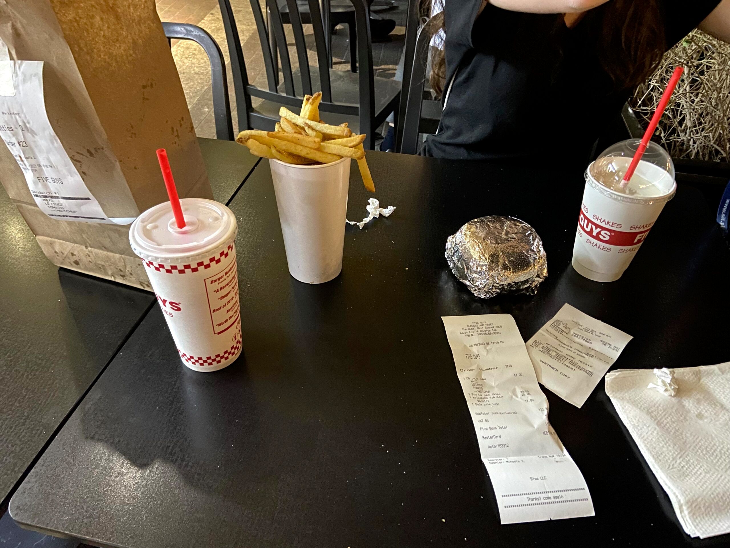 Posiłek w five guys w dubaju: frytki, burger, napoje i rachunki na czarnym stole.