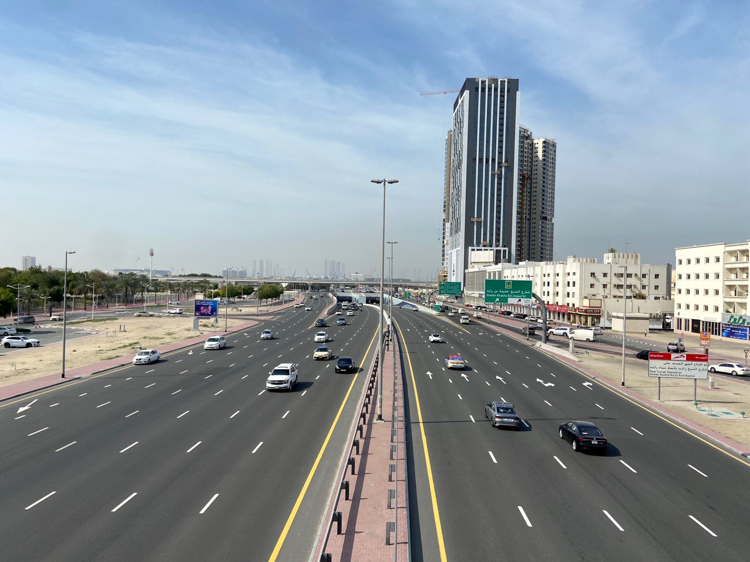 Szeroka autostrada w dubaju z ruchem samochodowym i wysokimi wieżowcami w tle.