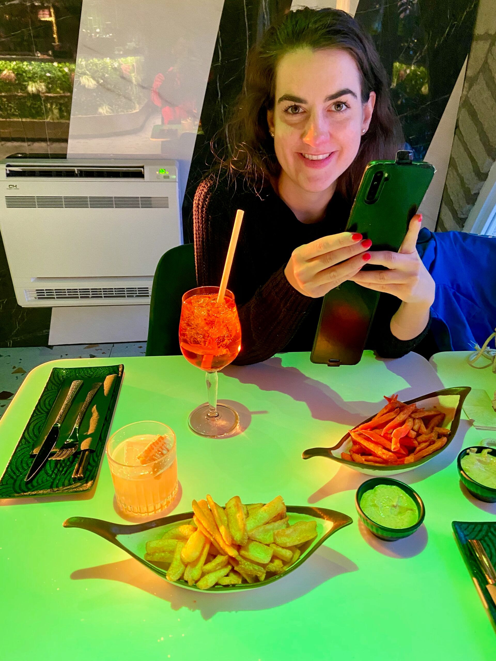 Woman in tallinn holding a green phone while dining at a table with fries and colorful drinks