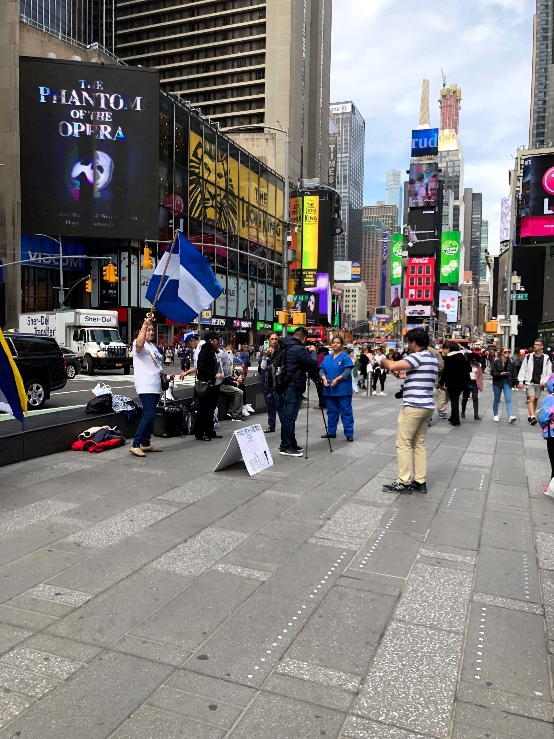 Times square w nowym jorku z tłumem ludzi, billboardami i osobą machającą niebiesko-białą flagą.