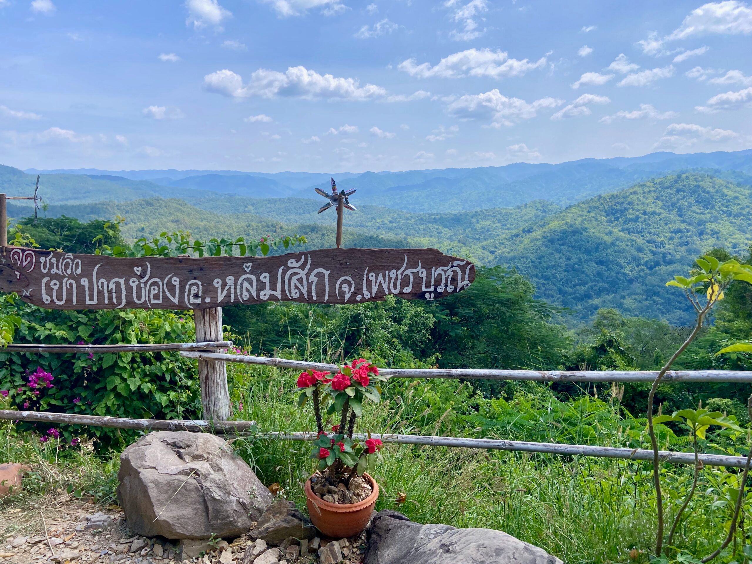Lom sak, thailand scenic viewpoint with a carved wooden sign, pink flowers, and layered green mountains under blue skies