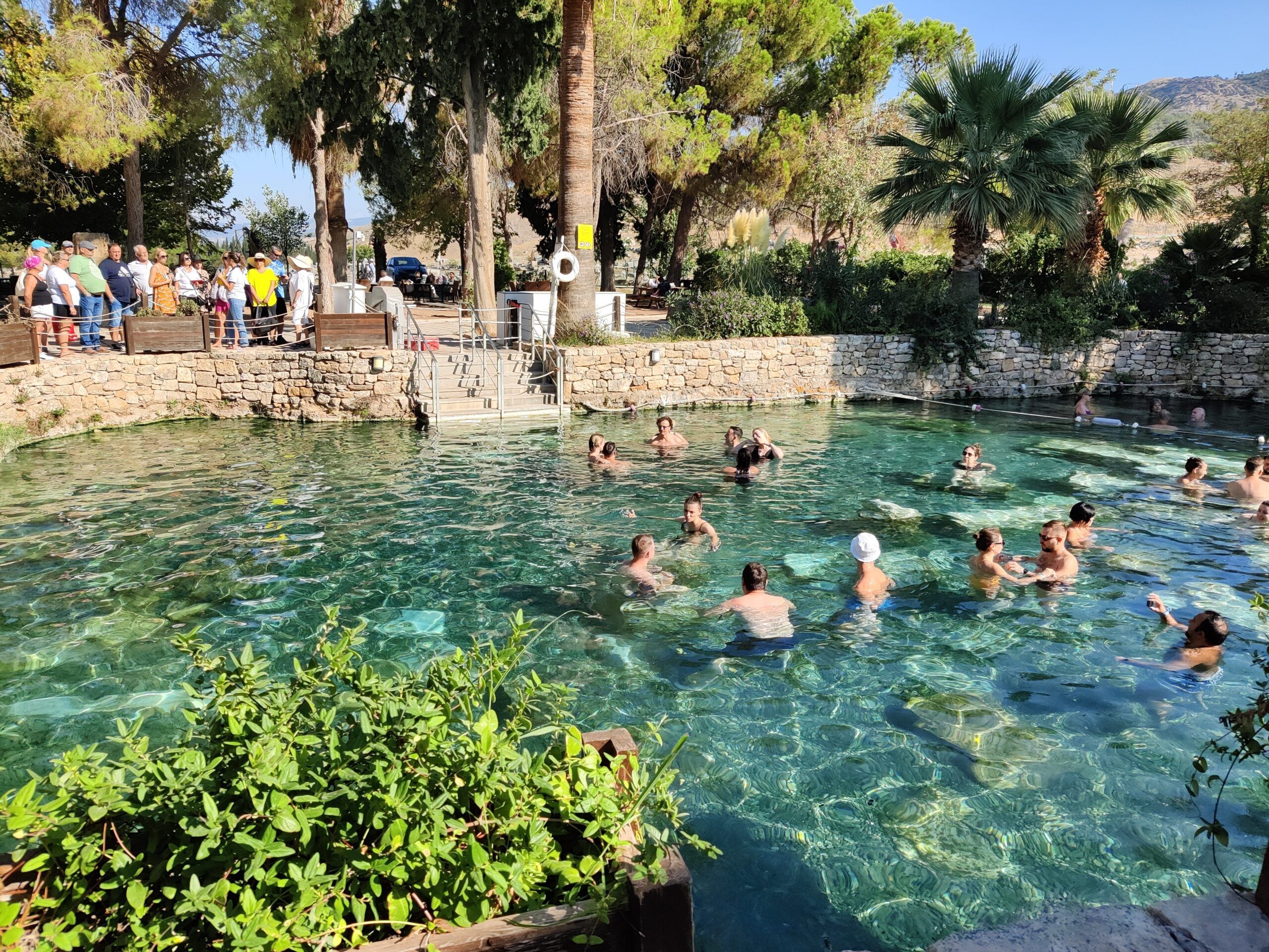 Pamukkale cleopatra antique pools w turcji, z ludźmi kąpiącymi się w krystalicznej wodzie otoczonej palmami.