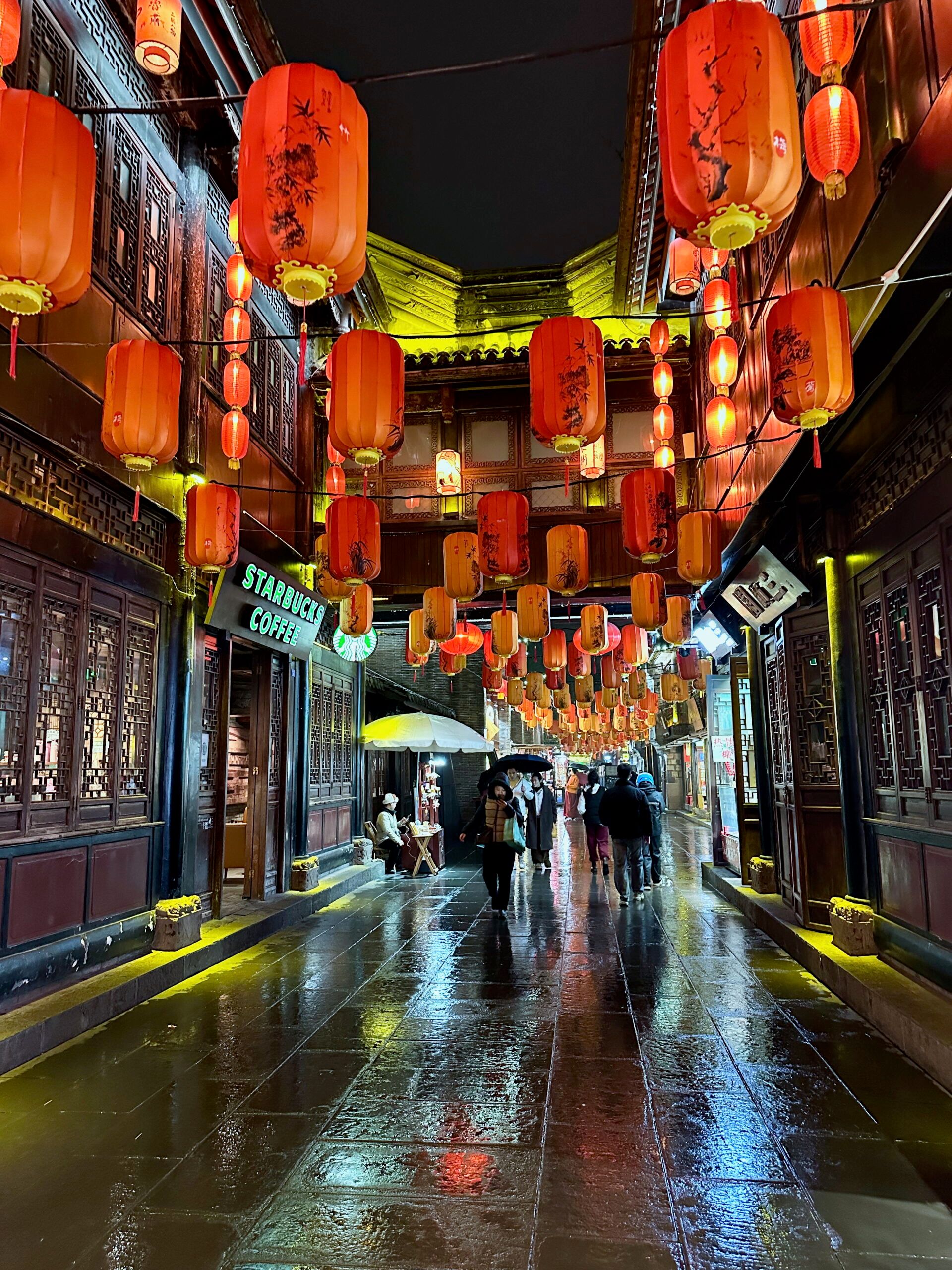 Chengdu jinli street nocą, z czerwonymi lampionami nad mokrą ulicą i spacerującymi ludźmi.