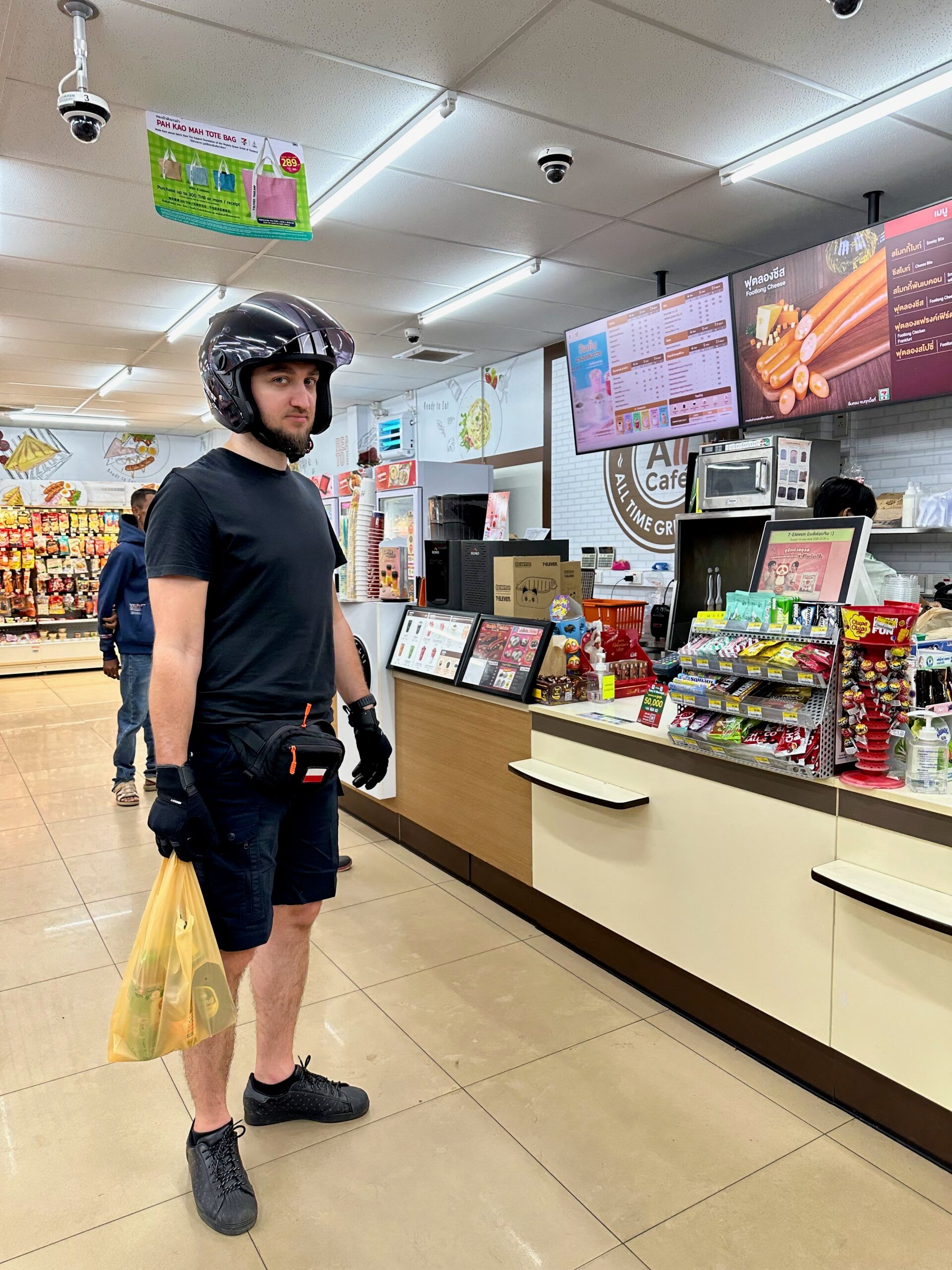 Mężczyzna w kasku stoi przy ladzie sklepu 7-eleven w phuket, tajlandia, z torbą zakupów.