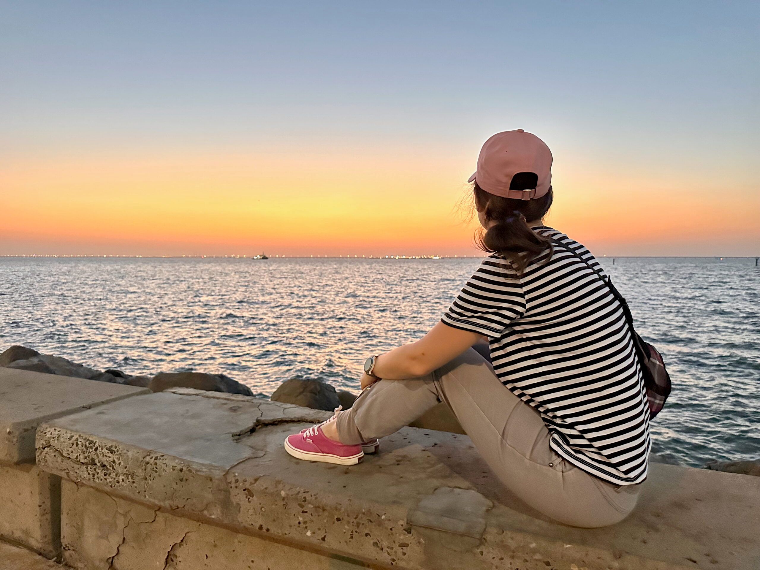 Woman in striped shirt and cap sitting by the sea at city souq sharq marina, kuwait, at sunset