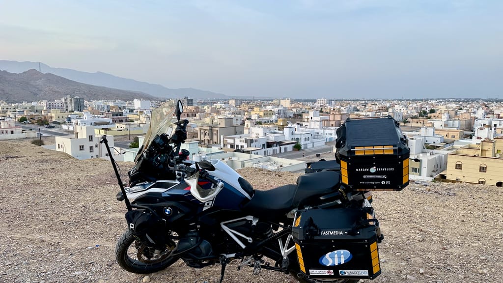 Motocykl turystyczny na wzgórzu nad miastem w omanie, z panoramicznym widokiem na zabudowę i góry.