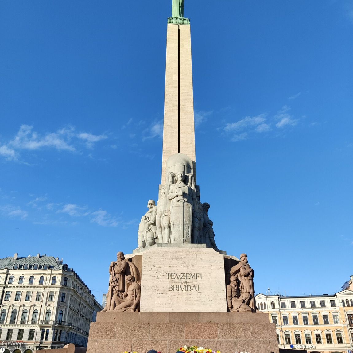 Pomnik wolności w rydze z ludźmi składającymi kwiaty u jego podstawy.