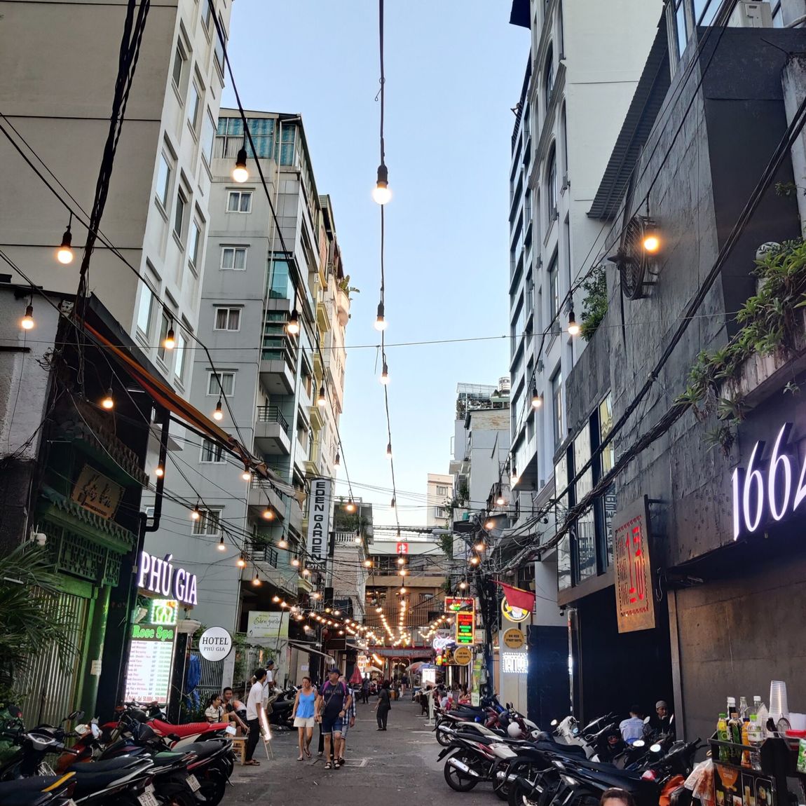 Ulica w ho chi minh z kawiarniami, motorami i wiszącymi lampkami między wysokimi budynkami.