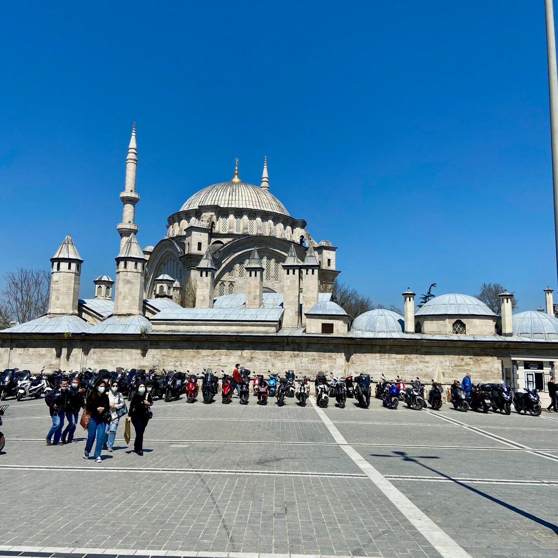 Meczet w stambule z kopułą i minaretami, przed nim zaparkowane motocykle i spacerujący ludzie.