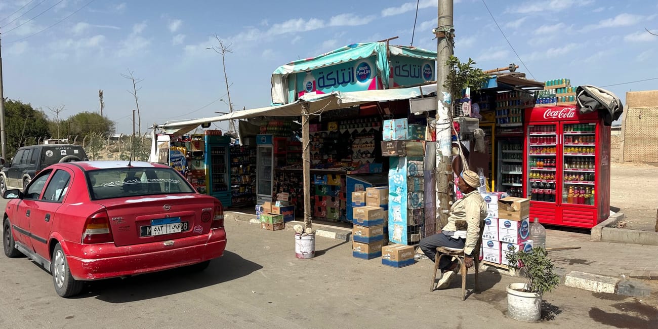 A roadside convenience shop in egypt with parked cars and a seated man under a blue sky