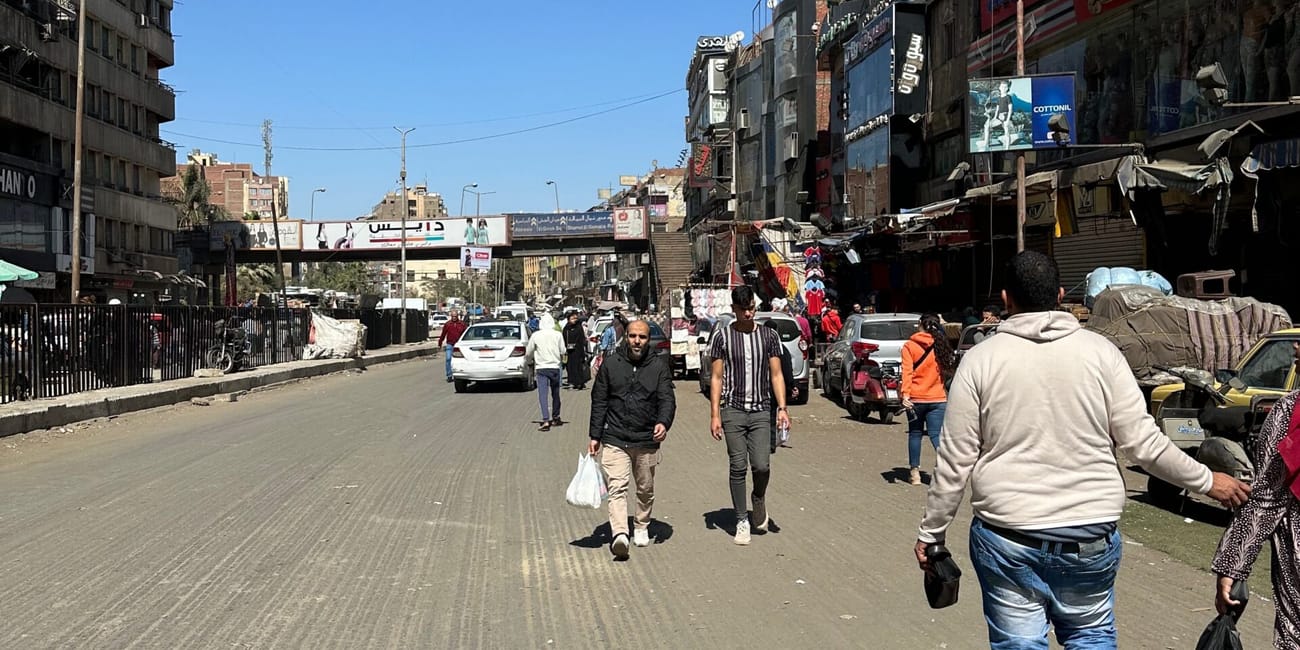 Busy street scene in egypt’s scams section with pedestrians, cars, and shops under a clear blue sky