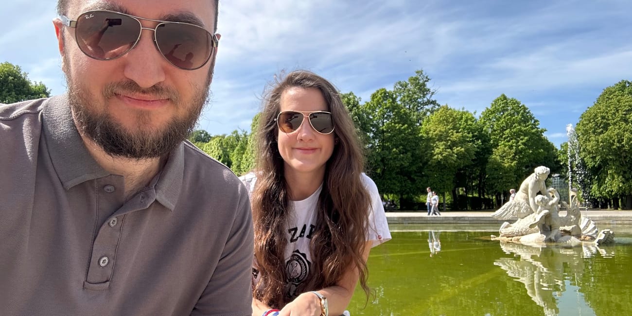 Dwoje turystów przy fontannie w wiedniu, z rzeźbą i parkiem w tle.