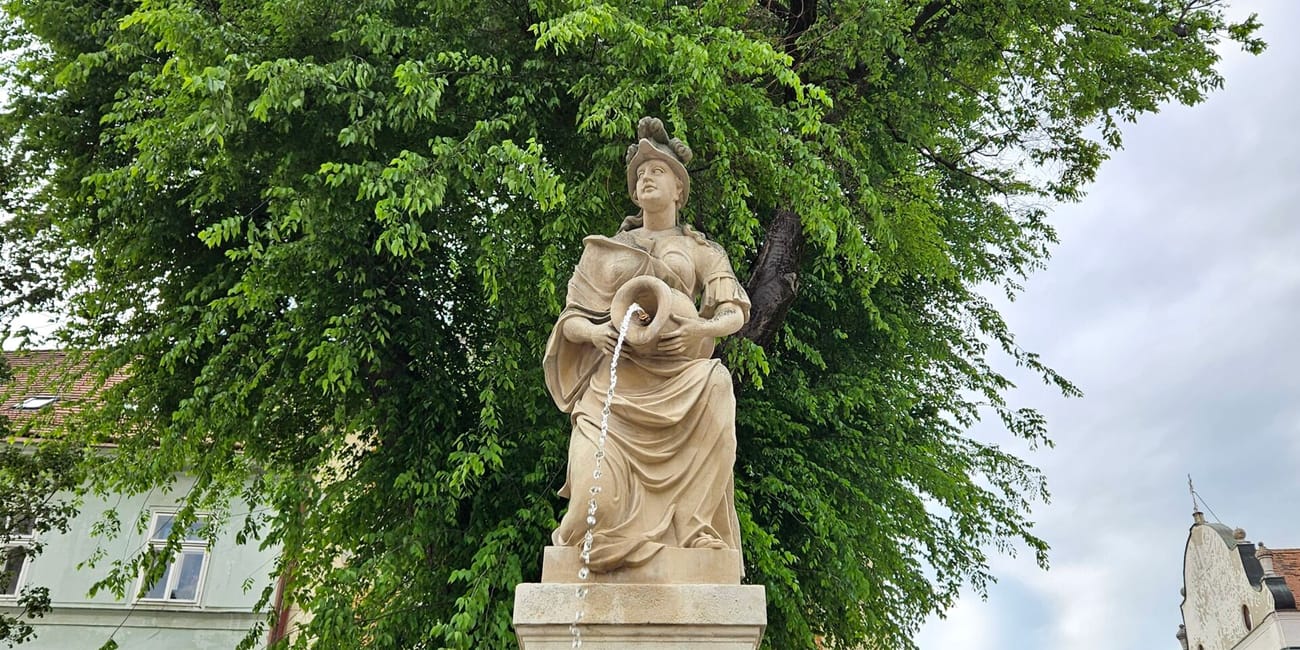 Fontanna z posągiem kobiety w zabytkowej części bratysławy, otoczona drzewami i kamienicami