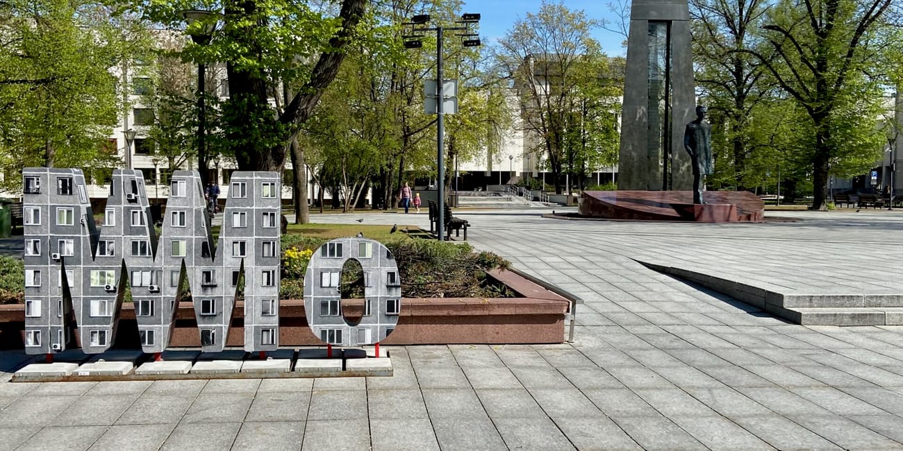 Plac z pomnikiem w wilnie, z zielonymi drzewami i dużym napisem mmo na pierwszym planie