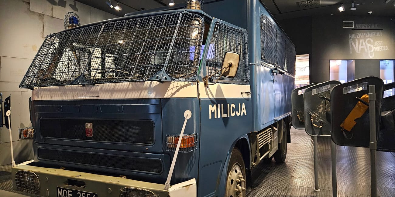 Niebieski samochód milicyjny w muzeum, z kratami ochronnymi i napisem milicja.