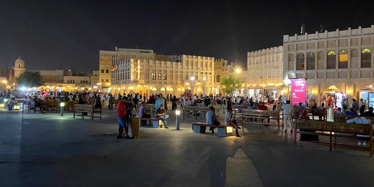 Nocny plac w dosze z tłumem ludzi, ławkami i oświetlonymi tradycyjnymi budynkami.