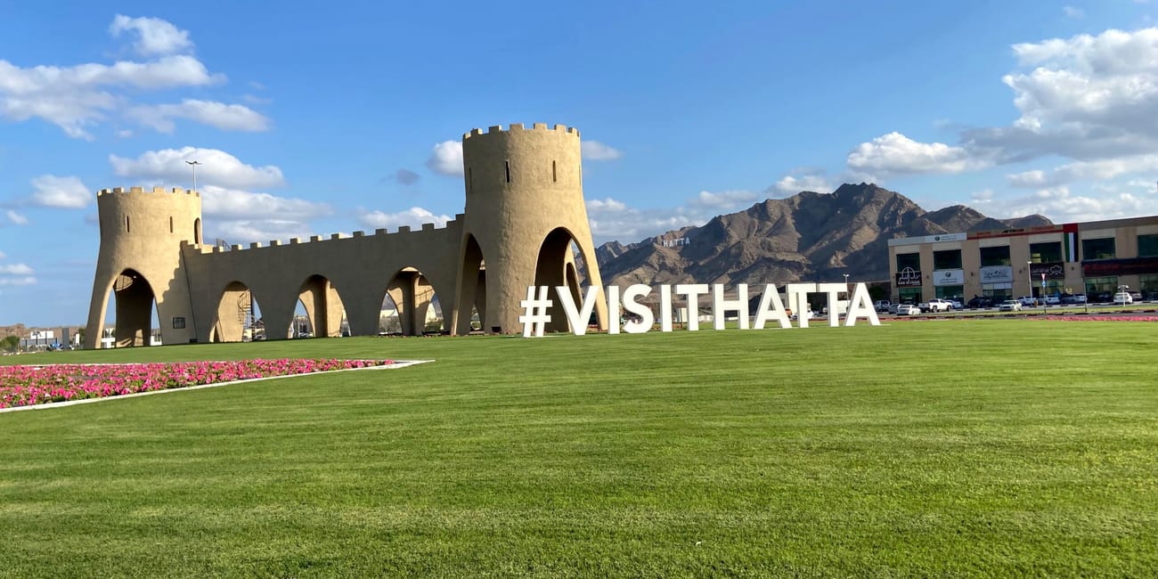 #visithatta w hatta, z wieżami, zielonym trawnikiem i górami w tle