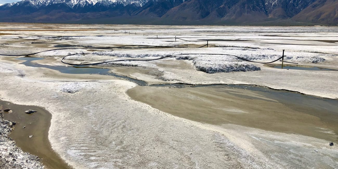 Rozległe solnisko w death valley z pasmem gór w tle и płytkimi strumieniami wody.