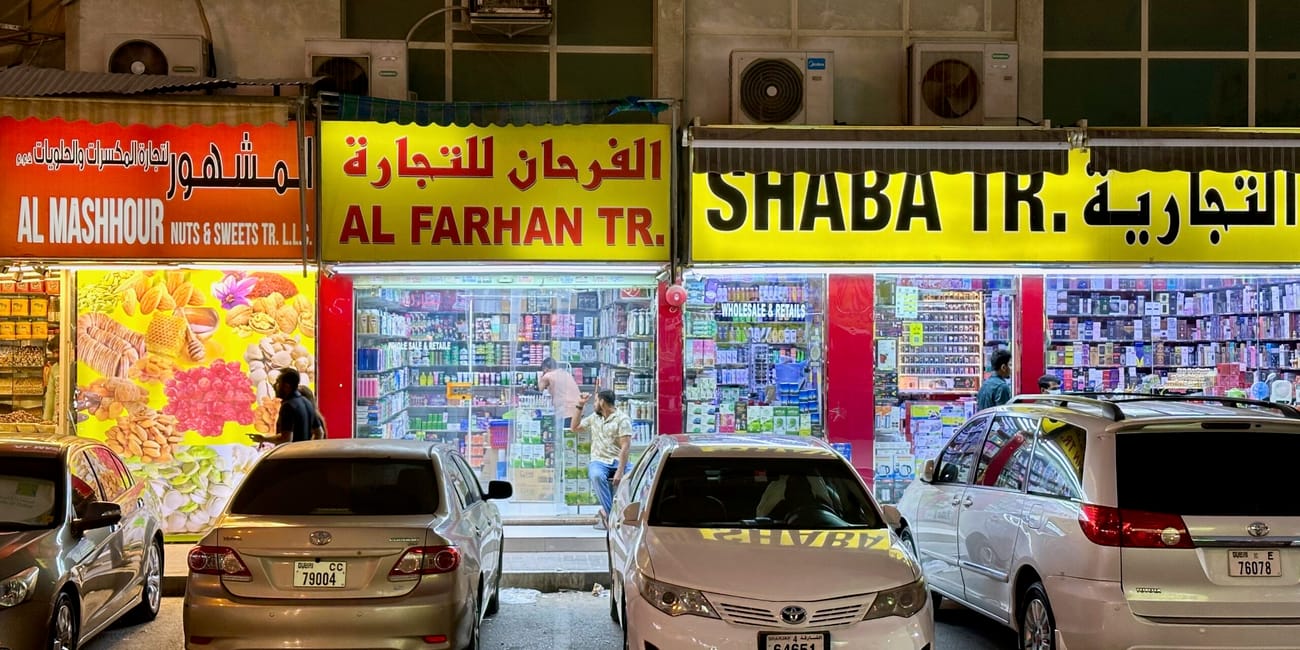 Sklepy przy al ghuwair market w sharjah nocą, z zaparkowanymi samochodami przed witrynami.