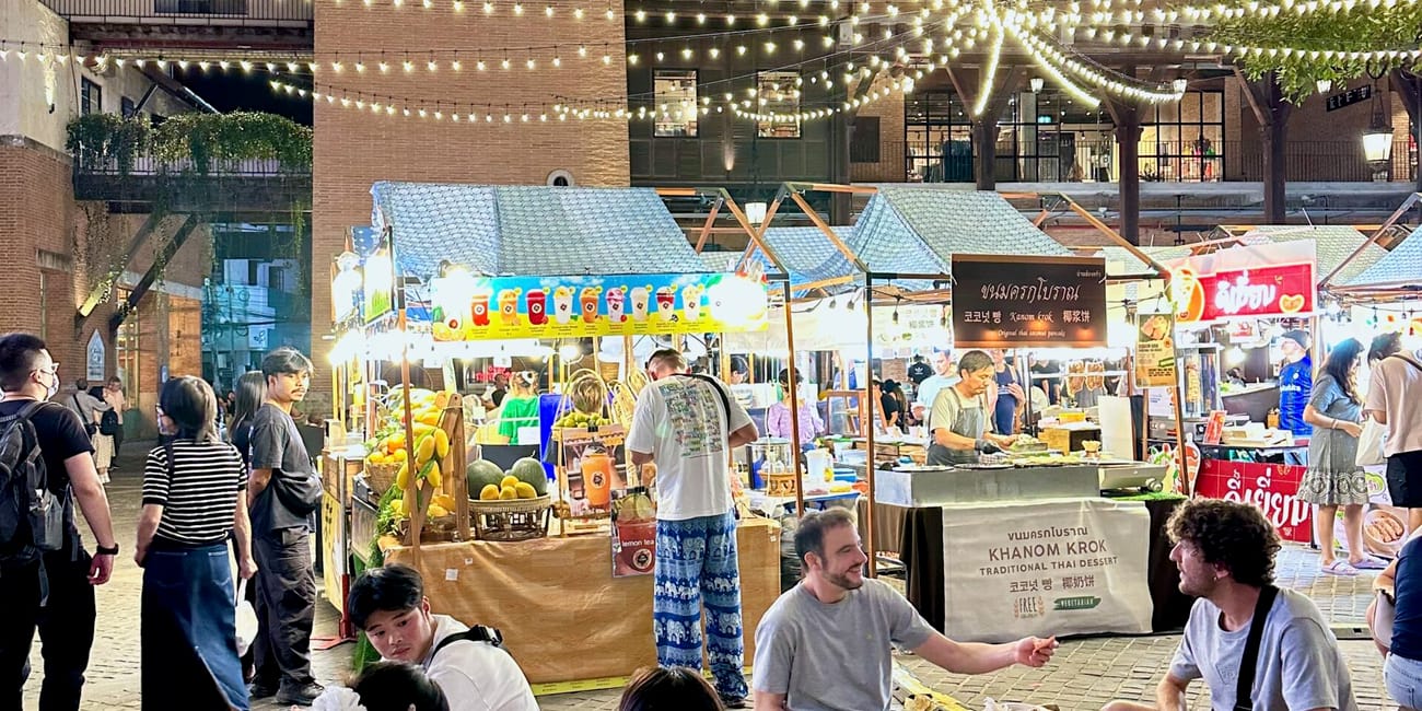 One nimman w chiang mai nocą, z podświetlonymi straganami i siedzącymi przy stoliku ludźmi.