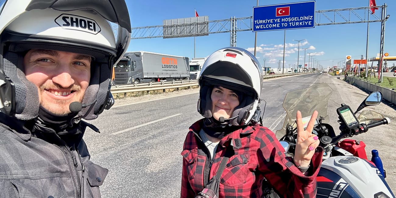 Dwoje motocyklistów przy znaku „welcome to türkiye” na drodze w turcji, z jednym motocyklem obok.