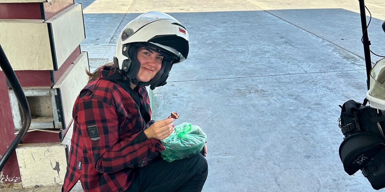 Motocyklowa ekspedycja na postoju przy stacji w iraku; kobieta w kasku je przekąskę.