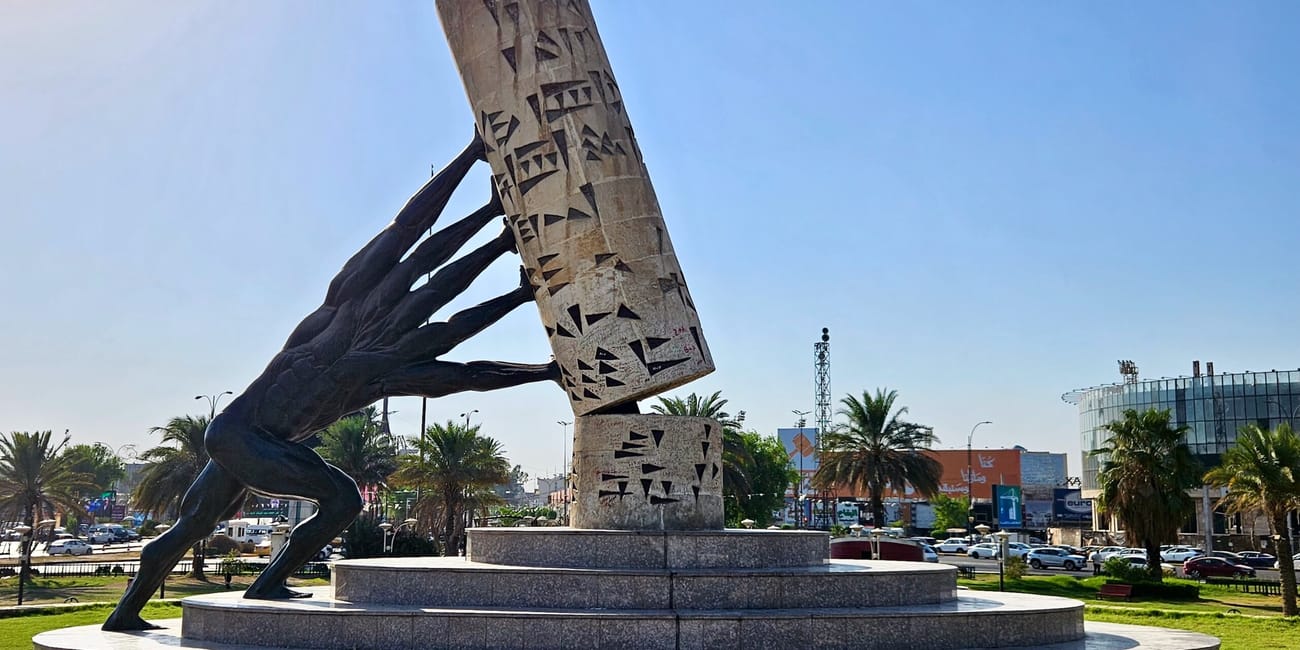 Monumentalna rzeźba kobiety dźwigającej popękaną płytę z mozaiką, na tle palm i ruchliwej ulicy w słońcu