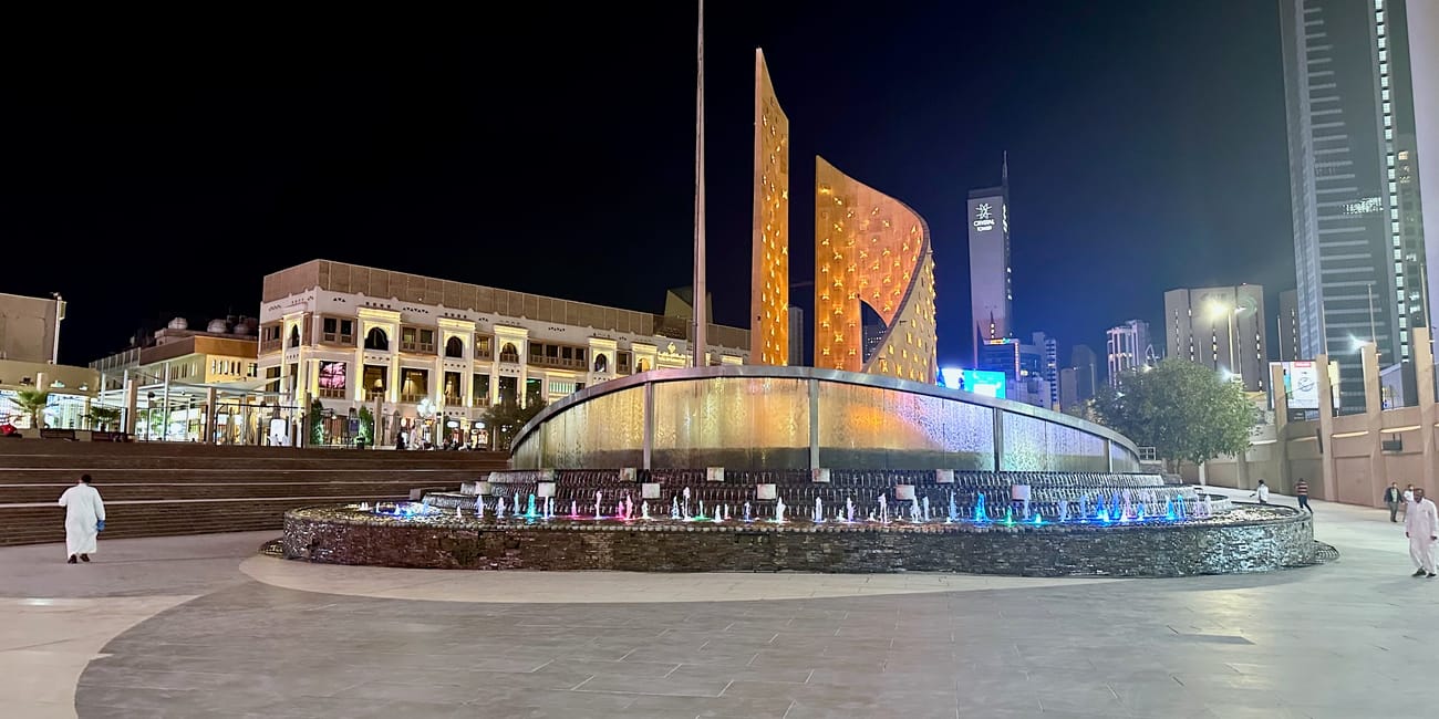 Kuwait city safat square at night with a lit fountain, flagpoles, and skyline buildings