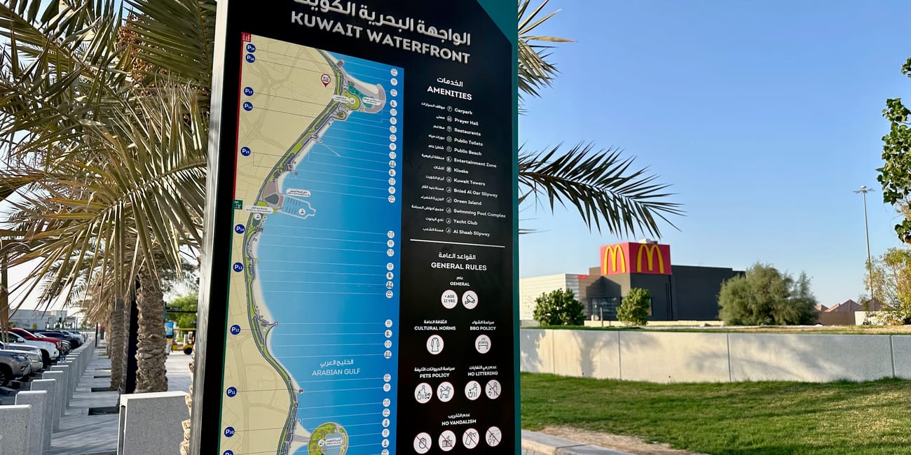 Kuwait city waterfront map sign with arabic and english text, palm trees, and a mcdonald’s storefront in the background