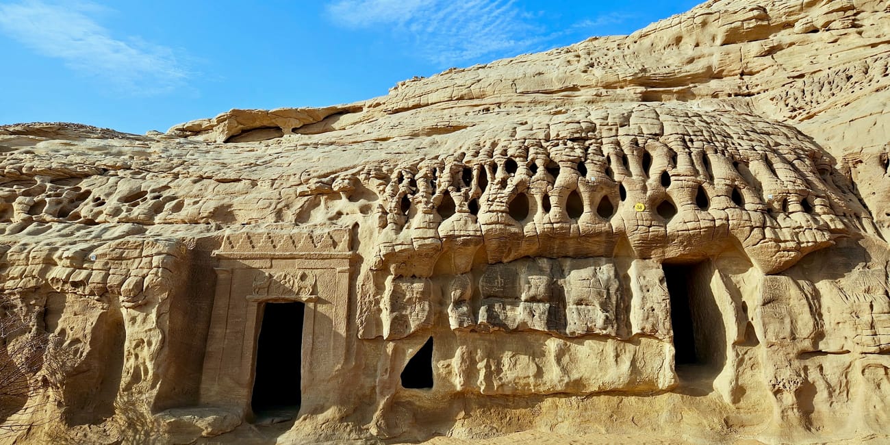 Saudi arabia, alula hegra tomb facade with arched doorways and carved rock openings under a blue sky