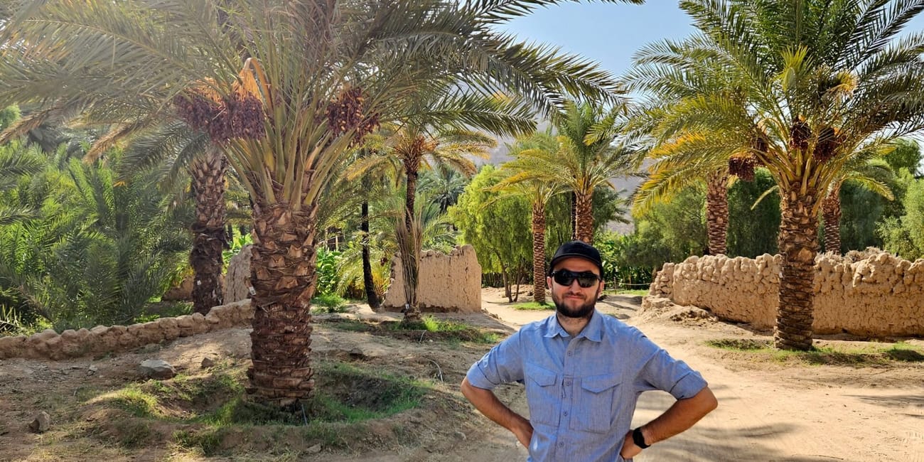 Man posing by al ula oasis near old town in saudi arabia, with palm trees and sandy paths behind him