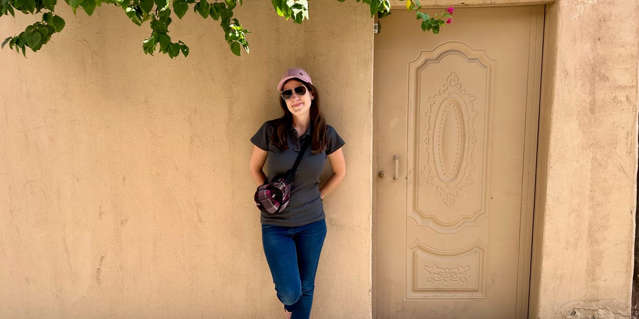 Woman standing in al ula old town, saudi arabia, beside an ornate beige door under hanging green leaves