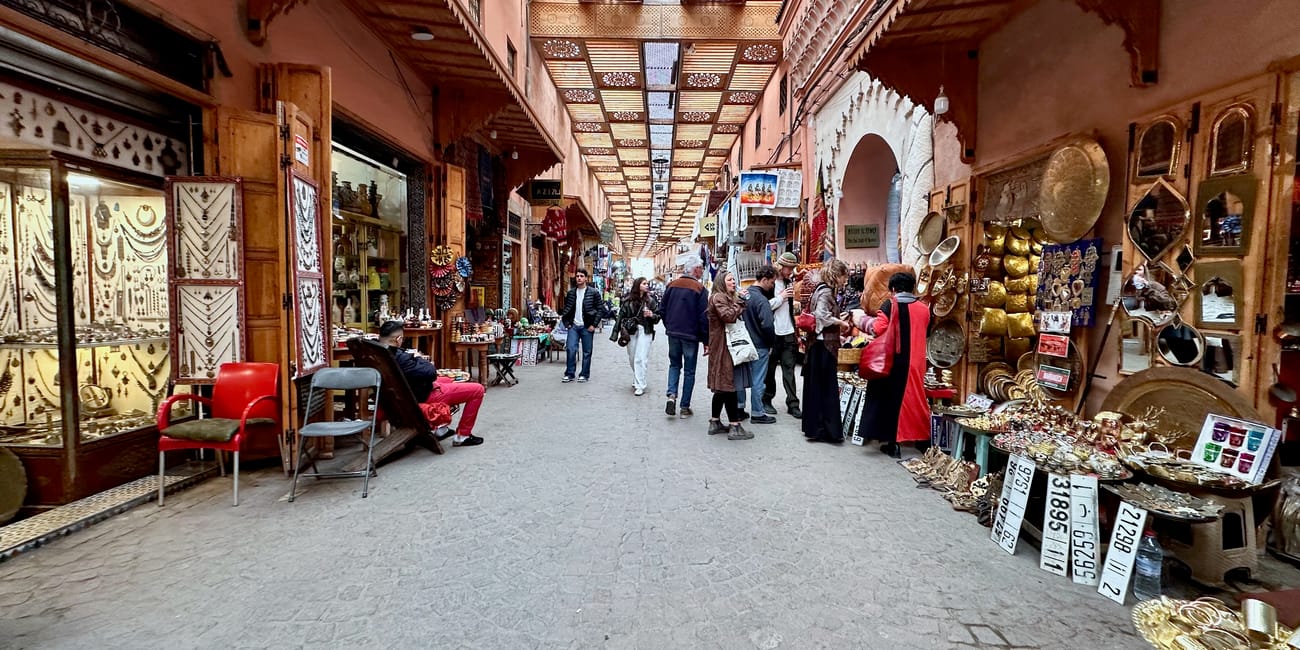 Souk semmarine in marrakech, morocco, with gold shops, hanging trays, and shoppers walking down a covered market corridor