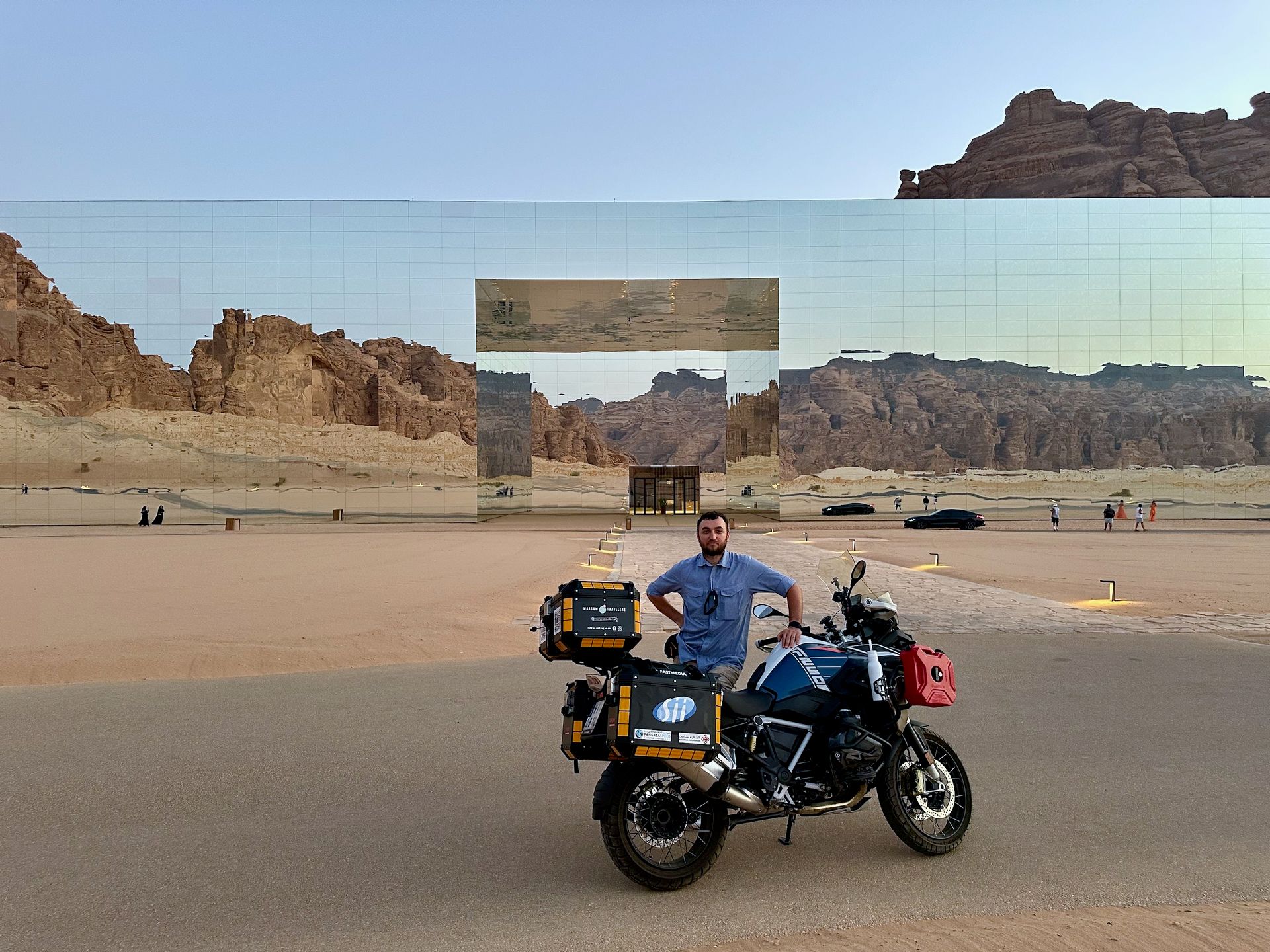 Motocyklista z wyprawowym motocyklem przed lustrzaną fasadą na pustyni w arabii saudyjskiej.