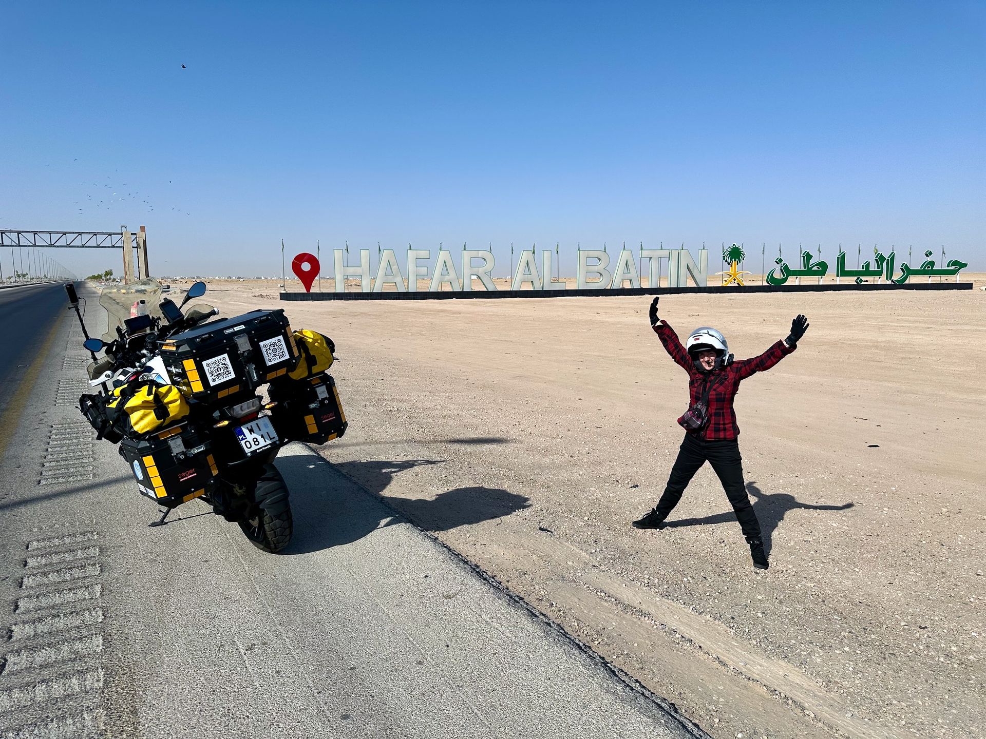 Motocykl wyprawowy przy napisie hafar al batin w arabii saudyjskiej, obok stojący motocyklista.