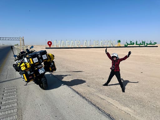 Motocykl wyprawowy przy napisie hafar al batin w arabii saudyjskiej, obok stojący motocyklista.