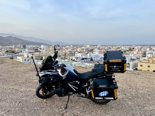 Motocykl turystyczny na wzgórzu nad miastem w omanie, z panoramicznym widokiem na zabudowę i góry.