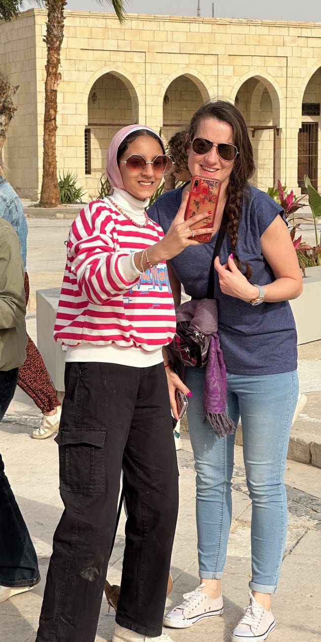 Two women posing for a selfie in egypt, with a stone building and arches behind them