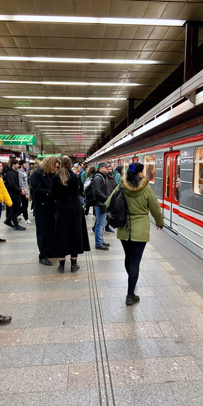 Peron metra w pradze z oczekującymi pasażerami i stojącym pociągiem.