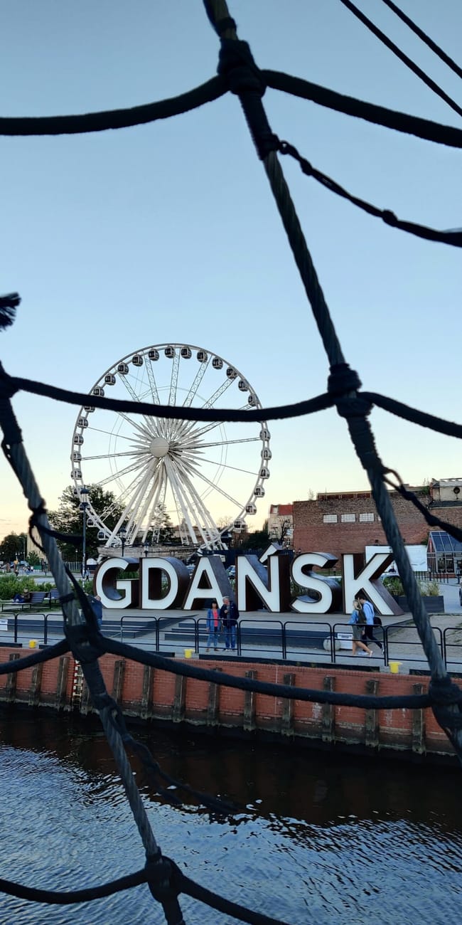 Koło widokowe i napis gdańsk nad motławą, widziane przez siatkę przy nabrzeżu.