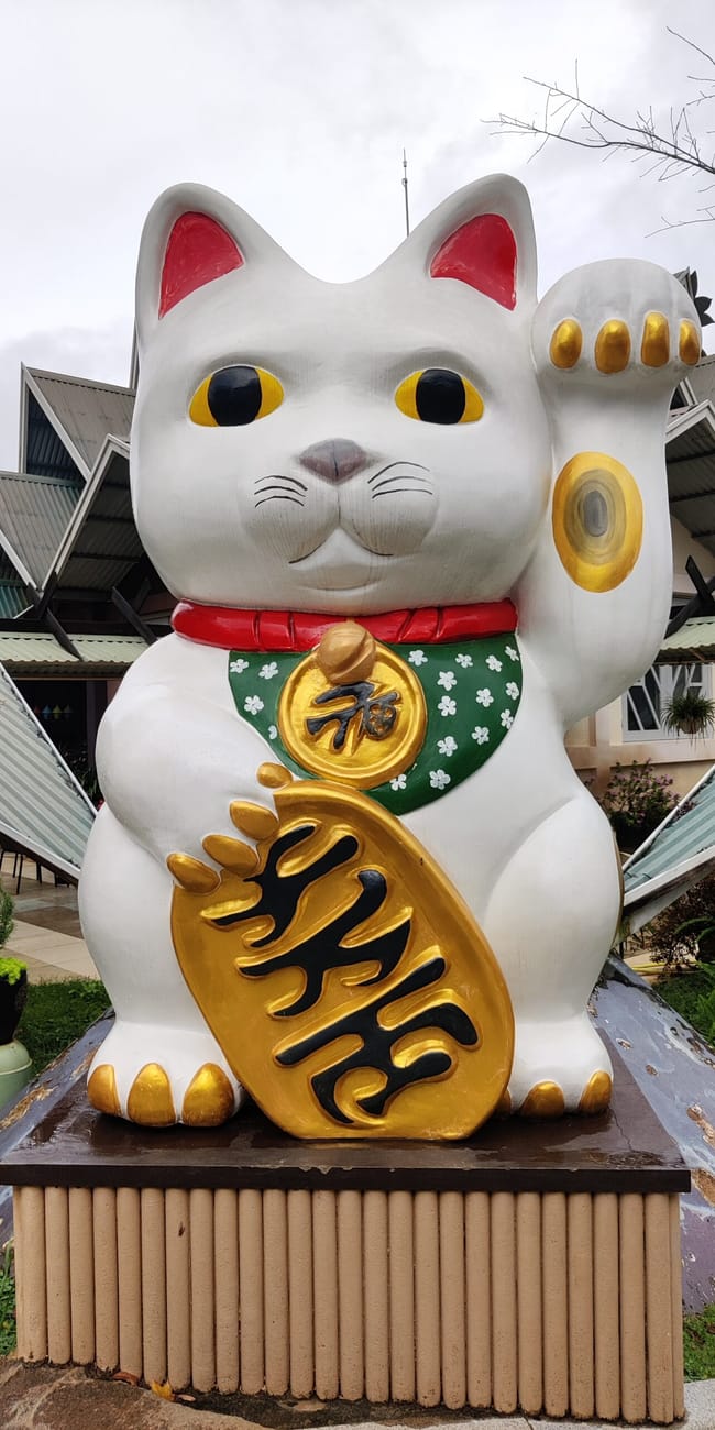 Rzeźba kota lucky cat w da lat kokoro cà phê, z podniesioną łapą i złotą monetą.